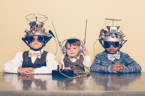 A young boy imagines reading minds of his two friends with a homemade science project. They are dressed in casual clothing, glasses and bow ties. They are serious and sitting at a table with helmets on their heads in front of a beige background. Retro styling.