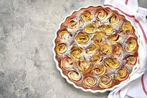 Homemade autumn pie with apple roses in a white baking form on a grey concrete or stone background.Top view.