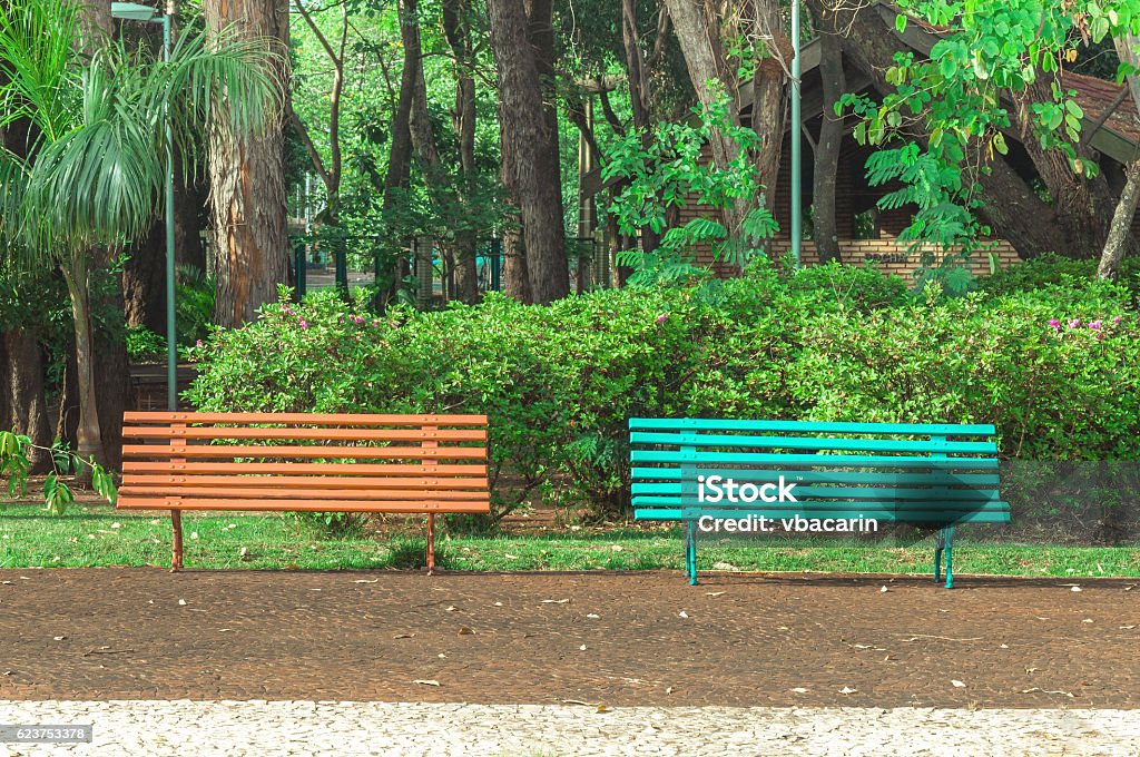 Two benches side by side on a square Two benches side by side on a square with trees and a beautiful green vegetation background. Orange and blue bench, very peaceful place to sit and relax with friends, choose one. Town Square Stock Photo