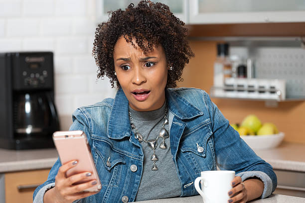 African American woman shocked what she sees on mobile phone Young African American woman on mobile phone with shocked look on her face, sitting in kitchen identity theft photos stock pictures, royalty-free photos & images