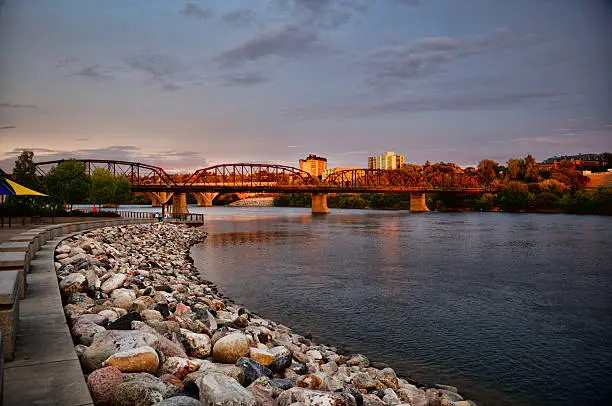 Sunset in Saskatoon at River Landing beside the South Saskatchewan River.  The sky is several shades of blue with the Traffic Bridge in the background.  Golden evening light is visible on the buildings in the background.  The River Landing promenade is visible to the left of the image.