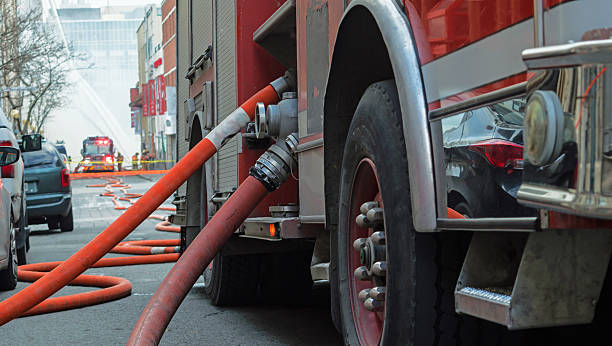 Fire truck hoses stretched out over city street to burning building with water flowing several stories high and team of firemen fighting fire in the background, selective focus on fire truck and fire fighting hose