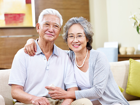 senior asian couple sitting on couch at home looking at camera smiling