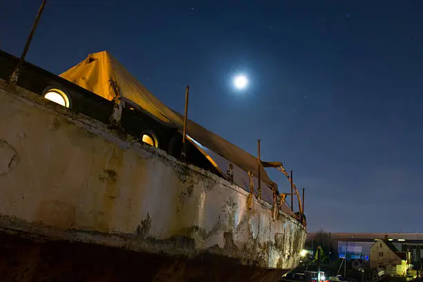 An old boat rests in dry dock with full moon overhead and industrial buildings behind, creating a nice atmosphere.