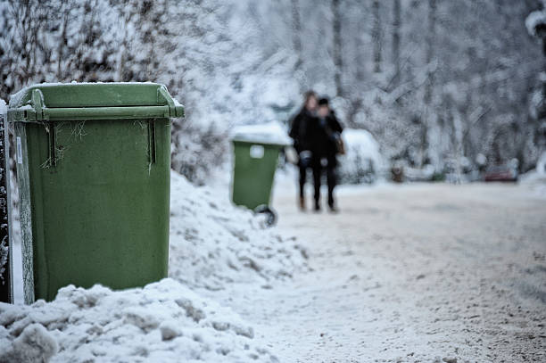 Waste bins on sidewalk in snow, pedestrians in background