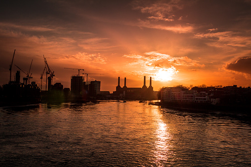 Wide angle image of the London skyline as seen from Vauxhall bridge. On the horizon we can see all kinds of construction equipment such as cranes, and blocks of flats or offices being built. The famous structure of Battersea power station can also be seen. The buildings are cast into silhouette as a dazzling sunset paints the sky golden orange, the colours reflected in the waters of the river Thames. Horizontal colour image with copy space.
