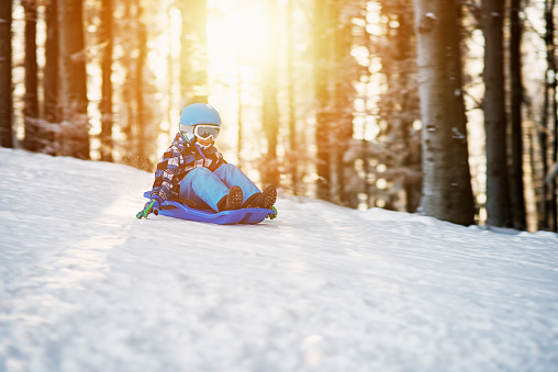 Little boy riding a sled in winter forest. Sunny winter day. The boy aged 6 is wearing a ski helmet and ski goggles.