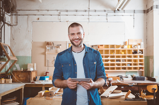 Small business. Young man in a carpentry workshop, holding a digital tablet in hands, smiling at camera.