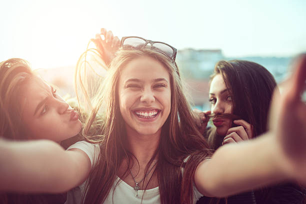 groupe de filles souriantes prenant drôle selfie en plein air au coucher du soleil - mobile phone group of people photographing teenager photos et images de collection