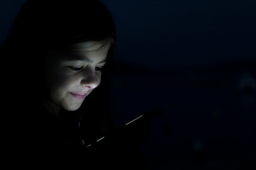 A young girl smiles happily as her face is lit up by the mobile phone screen as she is texting her friend in the dark.