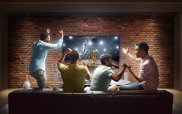 Students watching Basketball game at home :biggrin:A group of young male friends are cheering while watching Basketball game at home. They are sitting on a sofa in the modern living room faced to a big TV set on the front wall. tv game stock pictures, royalty-free photos & images