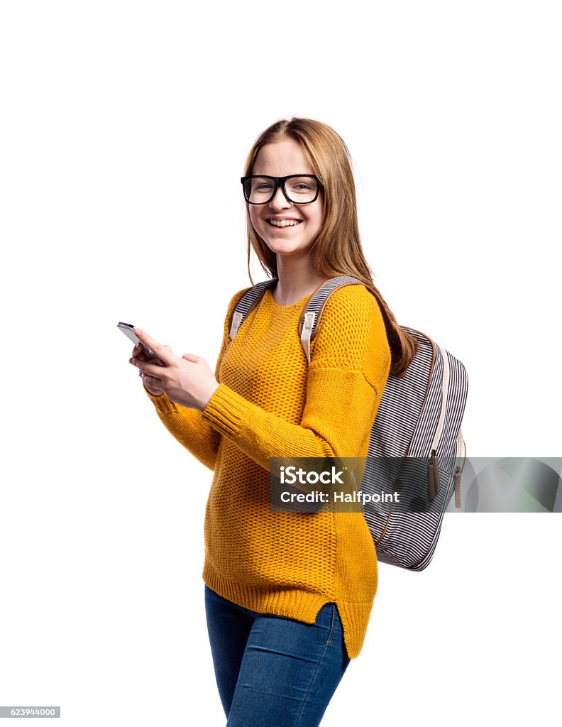 Girl in yellow sweater, holding smartphone, taking selfie, isola Teenage girl in jeans, mustard yellow sweater, with backpack on back. Young woman holding smart phone, taking selfie. Studio shot on white background, isolated. Teenager Stock Photo