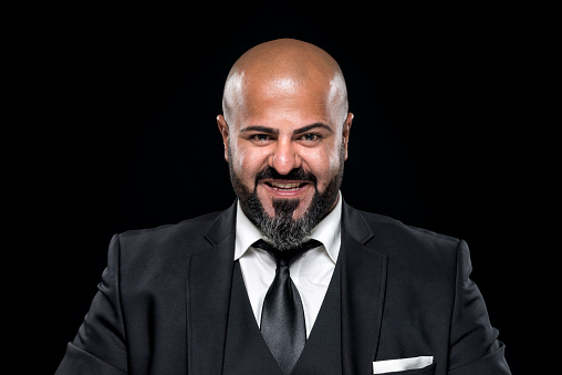 Bald businessman with black beard looking happy at camera. He is wearing an elegant black suit, a white button-down shirt and a tie. Studio shot in front of a black background.