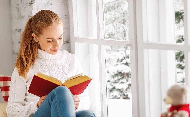 istock Happy young woman reading a book by window in winter 624009666