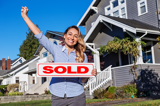 Photo of a cheerful young Hispanic businesswoman/Real Estate Agent with a red SOLD sign, standing in front of a row of suburban houses.