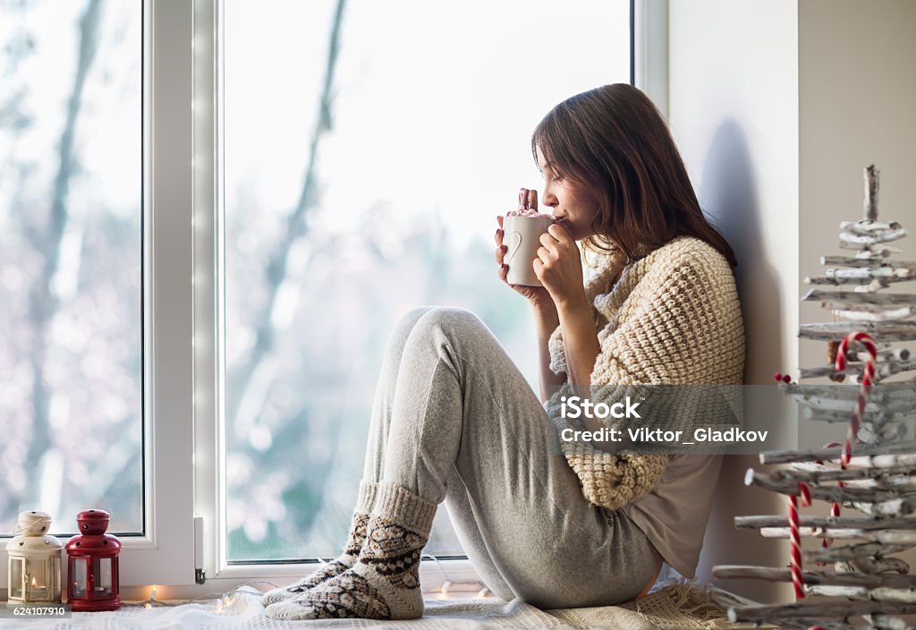 Young beautiful woman drinking hot coffee sitting on window sill Young beautiful woman drinking hot coffee sitting on window sill in christmas decorated home. Holiday concept Christmas Stock Photo