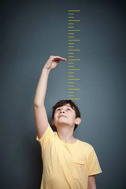 A cute boy is holding his arm up and showing his height on a wall scale.