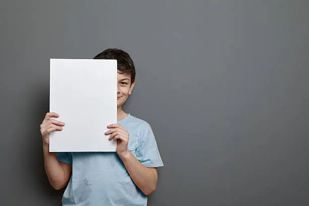 Cute child is holding a blank page, picture, sign or homework for copy space and hiding half of his face.
