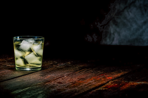 Drinking glass with ice cubes and yellow liquid on the dark wooden surface.