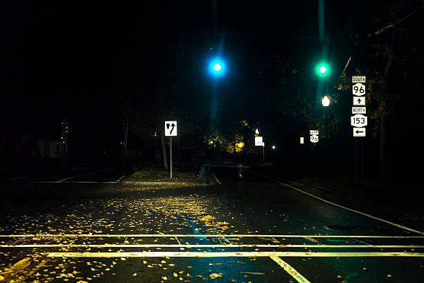 Late night lens flare traffic lights are green at a deserted, tranquil, quiet, stoplight controlled, village road intersection where drivers leave a residential district and enter the local business district of Pittsford, a suburban town near Rochester, NY in rural western New York State, USA. Driver POV perspective through a car windshield with the headlight high beams brightly illuminating the autumn leaf strewn intersection and foreground pedestrian crosswalk.