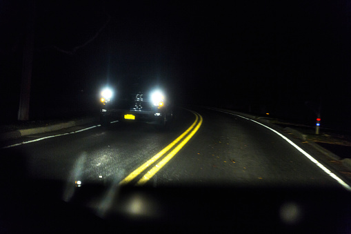 Driver POV perspective looking through the windshield of a car traveling on a slightly motion blurred rural highway blind curve. The road is strewn with blowing leaves on a very dark autumn night. A large SUV or pickup truck with bright headlights approaches from the opposite direction. NOTE: This image has some grain and a bit of noise.
