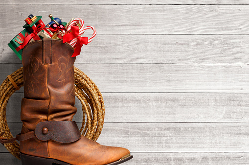 A lasso and a cowboy boot filled with Christmas goodies sit in front of whitewashed boards.  The Christmas items that spill out of the top of the boot include candy canes, a small train, Christmas ornaments, and several small presents. The negative space created by the boards provides ample room for copy and text.