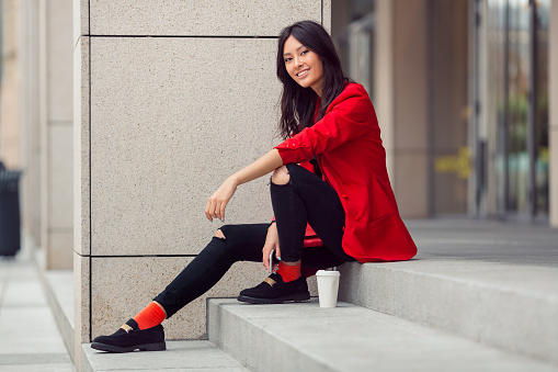 Happy asian businesswoman smiling in the city. Happy lovely and beautiful mixed race Asian Caucasian young woman in casual red suite outdoor sitting on steps of building with coffee cup. Next generation