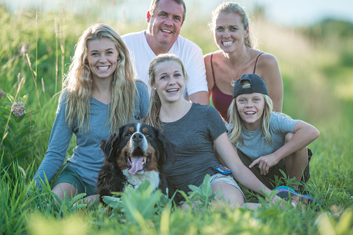 A family is sitting outside in a grassy field and are smiling while looking at the camera. Their dog is sitting with them happily.