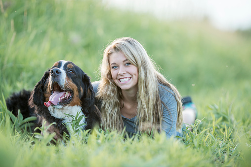 A teenage girl is lying in a grassy field with her pet dog. She is smiling and looking at the camera.