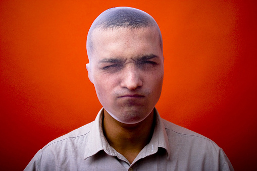 Angry young man with his head covered by a transparent stocking. Isolated on orange background, horizontal format.