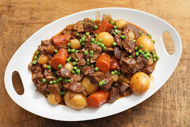 An overhead horizontal photograph of a white serving platter full of savory beef stew on a grungy wooden background.