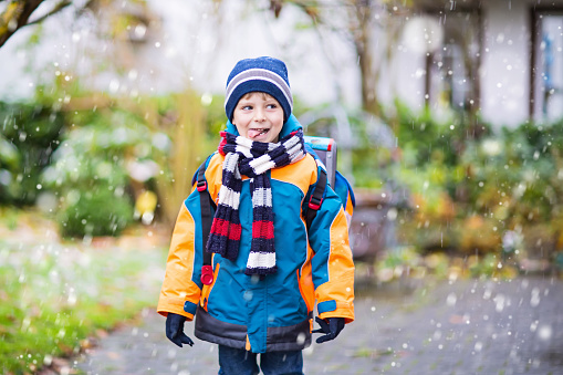 Little school kid boy of elementary class walking to school during snowfall. Happy child having fun and playing with first snow. Student with backpack in colorful winter clothes catching snowflakes.