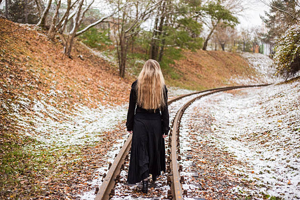 Woman in long black dress and hat walking on railway tracks. Winter. Cold weather. Travel alone.