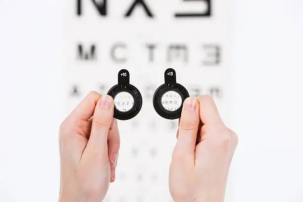 Bucharest, Romania - June 18,2016 : Closeup of hands holding longsightedness lenses in front of a blurry eye chart - isolated on white.
