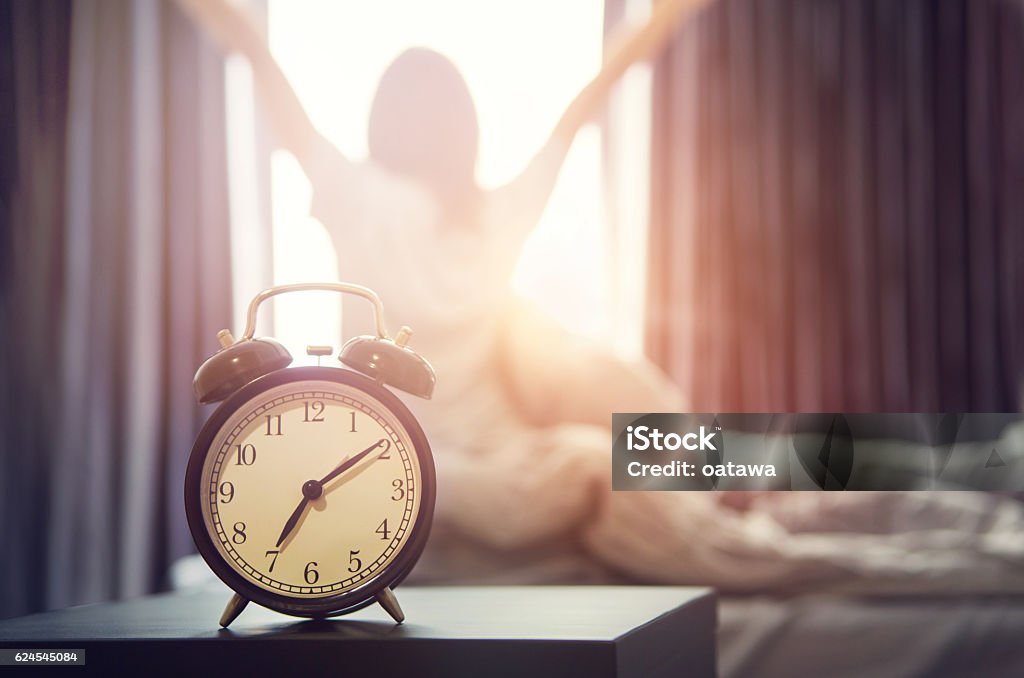 alarm clock having a good day in morning. Closeup alarm clock having a good day with background happy woman stretching in bed after waking up, sunlight in morning. Morning Stock Photo