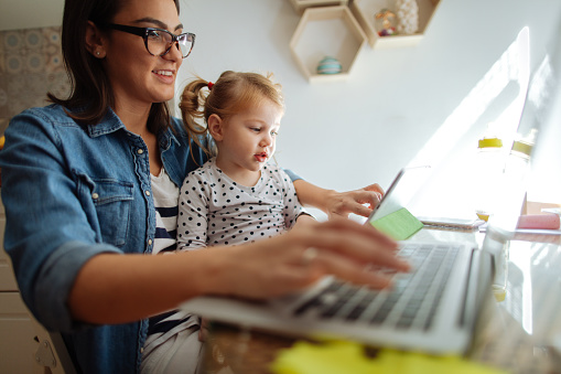 Photo of a little girl watching cartoons on a digital tablet, while her mom is working on a computer from their home