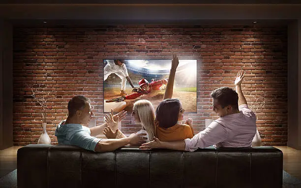 :biggrin:Two couples are cheering while watching Baseball game at home. They are sitting on a sofa in the modern living room faced to a big TV set on the front wall.