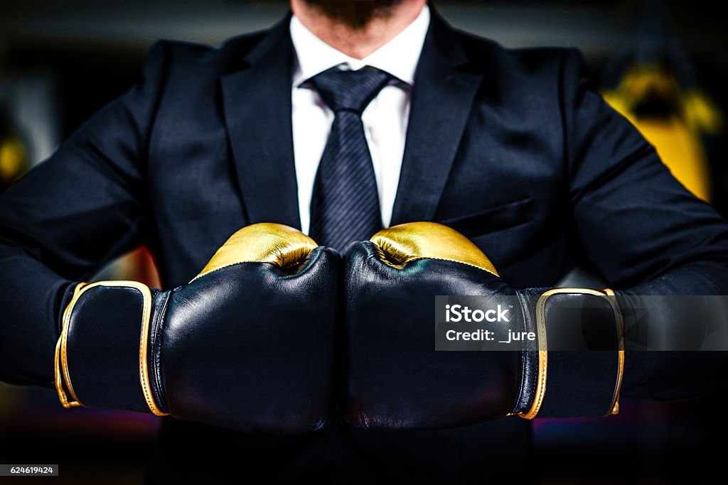 Businessman with boxing gloves is ready for corporate battle. Businessman with boxing gloves is ready for corporate battle. Man in suit, shirt and a tie is holding combat gloves together. Shot in a boxing gym, concept of relentless struggle and success. Boxing - Sport Stock Photo