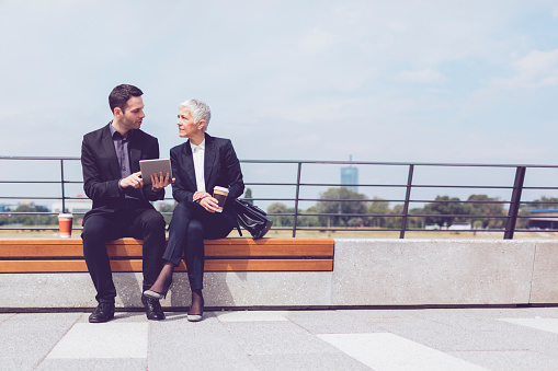 Businesspeople sitting on bench near fence by the riverside. They are having meeting out of office. Drinking coffee and working. Businessman holding digital tablet and talking to his colleague, mature businesswoman.