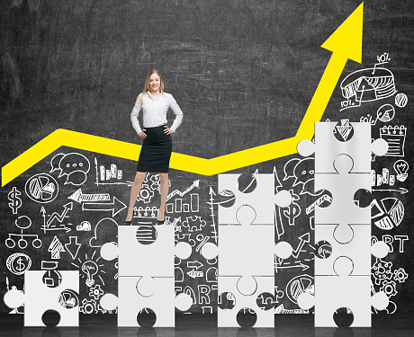 Front view of businesswoman standing on white puzzle pieces and near blackboard with yellow graph and business icons. Concept of success. Mock up