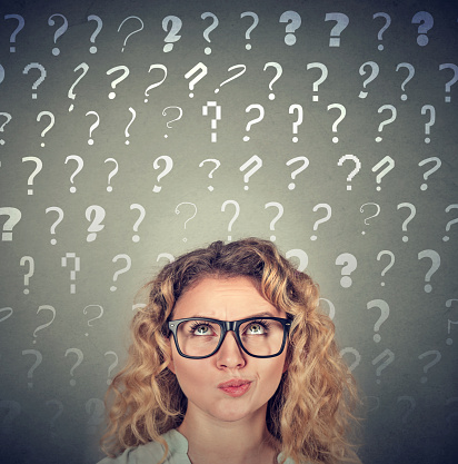 Thinking business woman with glasses looking up at many questions marks above head isolated on gray wall background