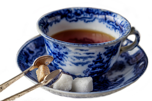 Antique blue and white china cup and saucer of black English breakfast tea with silver tongs for white and brown sugar cubes. Isolated on white background with copy space.