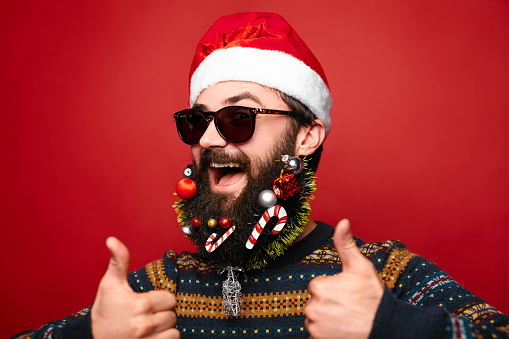 Happy modern Santa Claus showing thumbs up over vivid red background. Close up face of young man with decorated beard and sunglasses wearing santa hat.