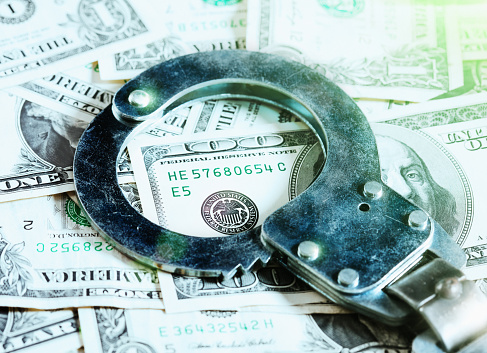 A pair of handcuffs rests on a US $100 bill among an abundance of dollars, symbolizing fraud, white collar crime, theft, moneylending and many other crimes.