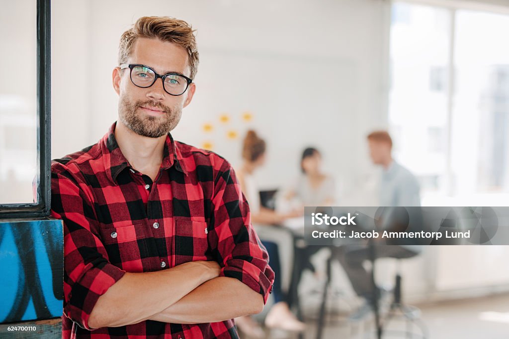 Portrait of positive businessman in office Portrait of a positive looking young business professional standing with his arms crossed with coworkers talking in the background. Men Stock Photo