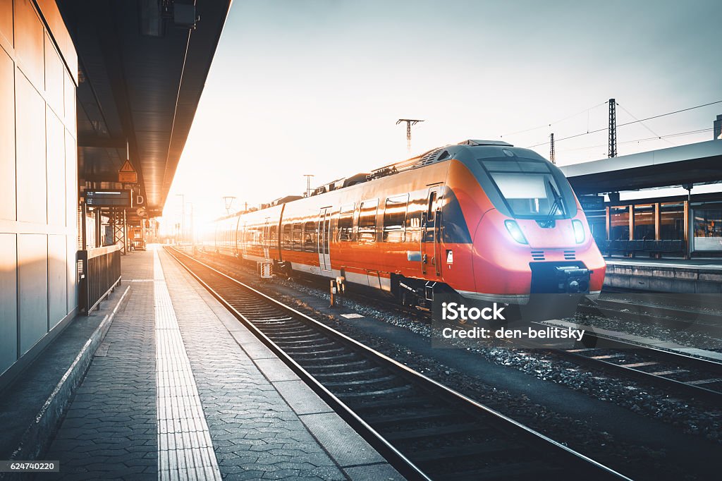 Modern high speed red commuter train at the railway station Modern high speed red commuter train at the railway station at sunset. Turning on train headlights. Railroad with vintage toning. Train at railway platform. Industrial landscape. Railway tourism Train - Vehicle Stock Photo