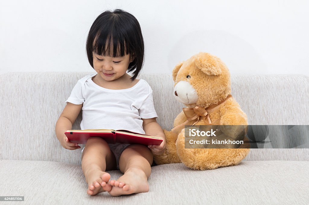 Asian Chinese little girl reading book with teddy bear Asian Chinese little girl reading book with teddy bear on the sofa in the living room. Child Stock Photo