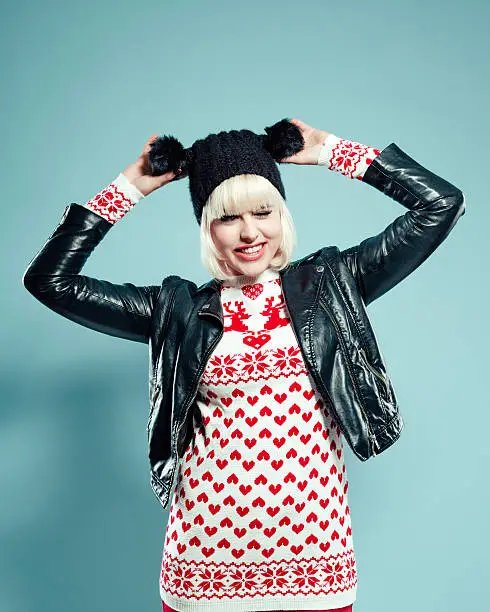 Portrait of excited blonde young woman wearing christmas sweater with reindeers, leather jacket and black bobble hat, holding bobbles and laughing at camera. Studio shot, turquoise background.