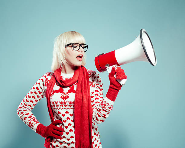 Blonde young woman wearing xmas sweater, holding megaphone Portrait of beautiful blonde young woman wearing christmas sweater, red scarf and gloves, talking into megaphone. Studio shot, turquoise background. christmas nerd sweater cardigan stock pictures, royalty-free photos & images