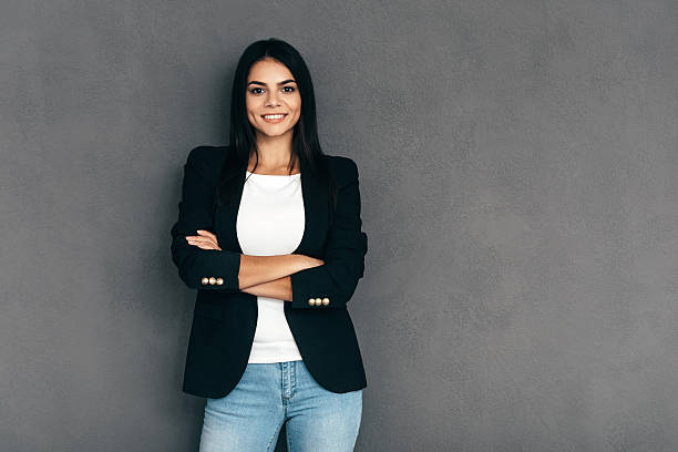 Confident and beautiful. Attractive young woman in smart casual wear keeping arms crossed and looking at camera with smile while standing against grey background smart casual stock pictures, royalty-free photos & images